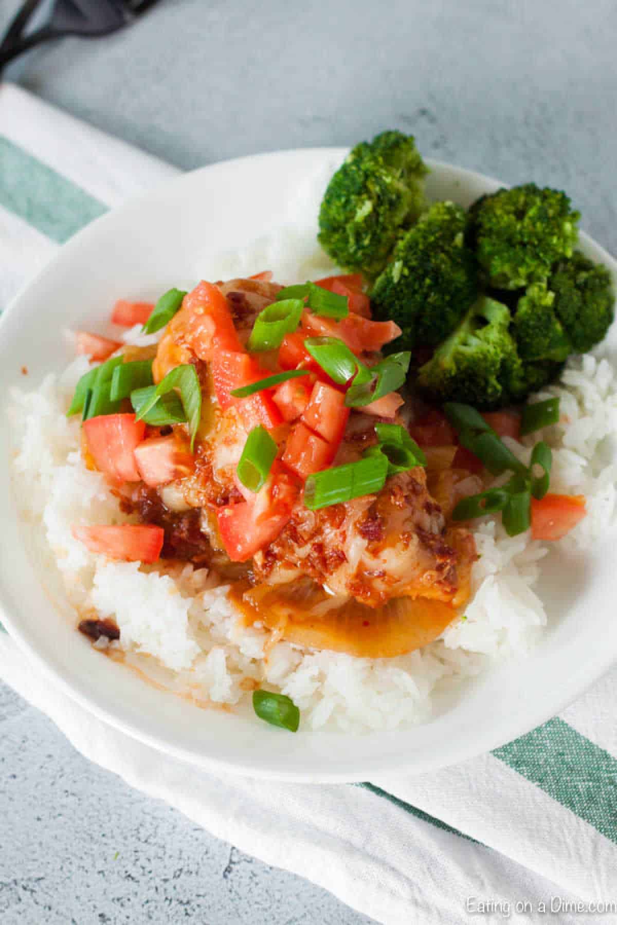 A plate of rice topped with crock pot Monterey chicken, diced tomatoes, green onions, and sauce. The dish is accompanied by a side of steamed broccoli. A striped green and white napkin is partially visible underneath the plate.