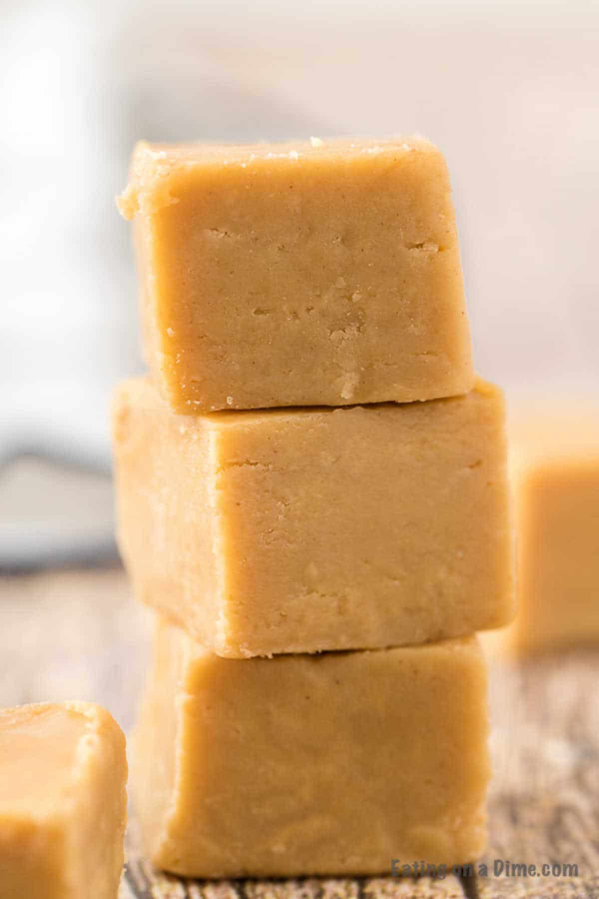 Three pieces of creamy, tan-colored 3 Ingredient Peanut Butter Fudge are stacked on top of each other against a blurred background, with one additional piece partially visible in the foreground. The fudge has a smooth texture with slight imperfections on the surface.