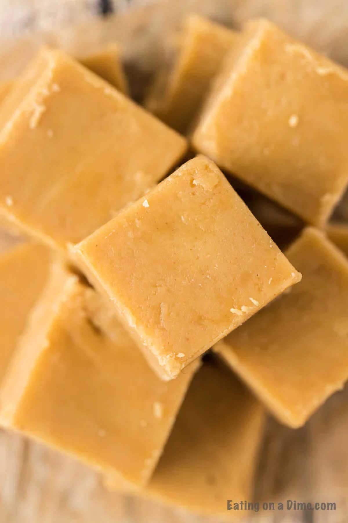 Close-up view of a stack of creamy, light brown 3 Ingredient Peanut Butter Fudge squares. The texture appears smooth and slightly crumbly along the edges. The background is softly out of focus, and there is a faint watermark at the bottom right that reads "Eating on a Dime.com.