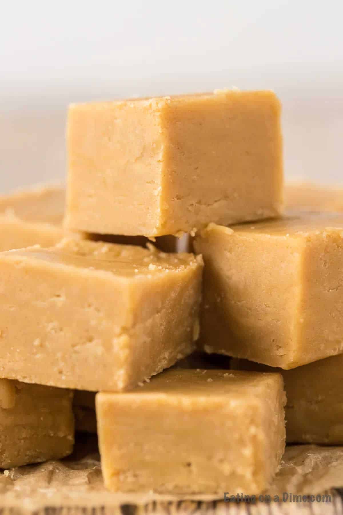 A close-up view of several square pieces of creamy 3 Ingredient Peanut Butter Fudge stacked unevenly. The fudge has a smooth texture with a light, tan color. The background is softly blurred, highlighting the rich, delectable appearance of the fudge squares.