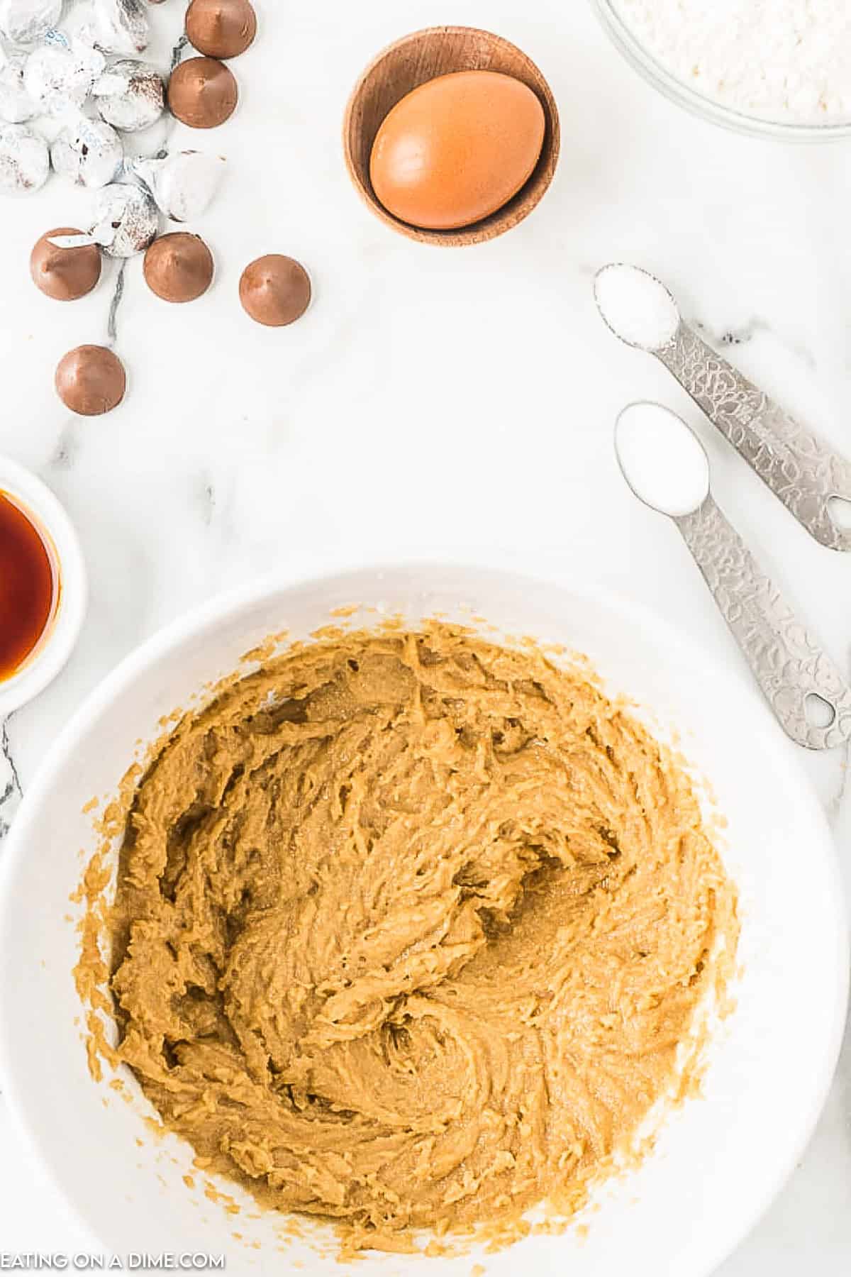 A white mixing bowl filled with cookie dough for peanut butter kiss cookies is on a countertop. Surrounding the bowl are several ingredients: an egg in a wooden bowl, flour in a glass bowl, Hershey's kisses, measuring spoons with salt and baking soda, and a small dish of vanilla extract.