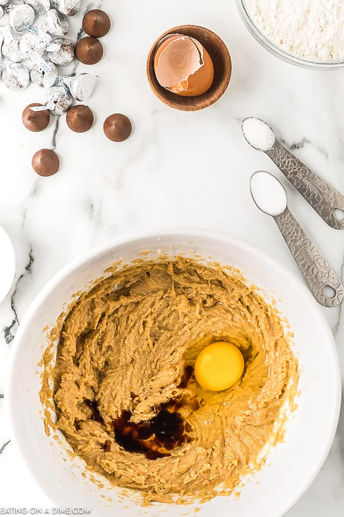 A mixing bowl contains an almost fully mixed batter with a cracked egg and vanilla extract on top. Surrounding the bowl are ingredients for peanut butter kiss cookies, including flour, chocolate candies, salt, and a cracked eggshell, along with measuring spoons and a small bowl.