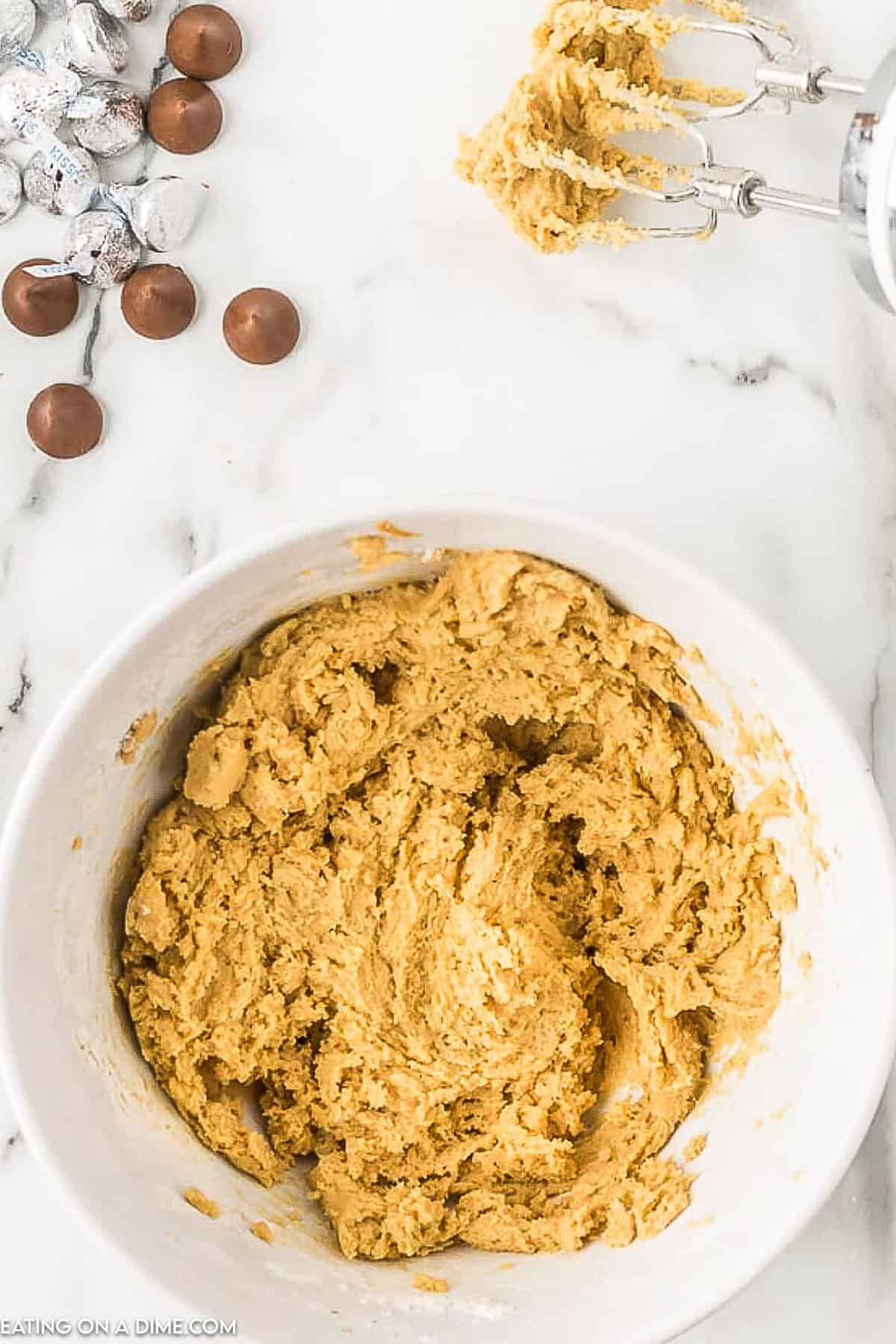 A white bowl filled with cookie dough is on a marble countertop. To the side, there are pieces of chocolate and a few chocolate kisses. An electric mixer with dough on the beaters is placed nearby, ready to create delicious peanut butter kiss cookies.