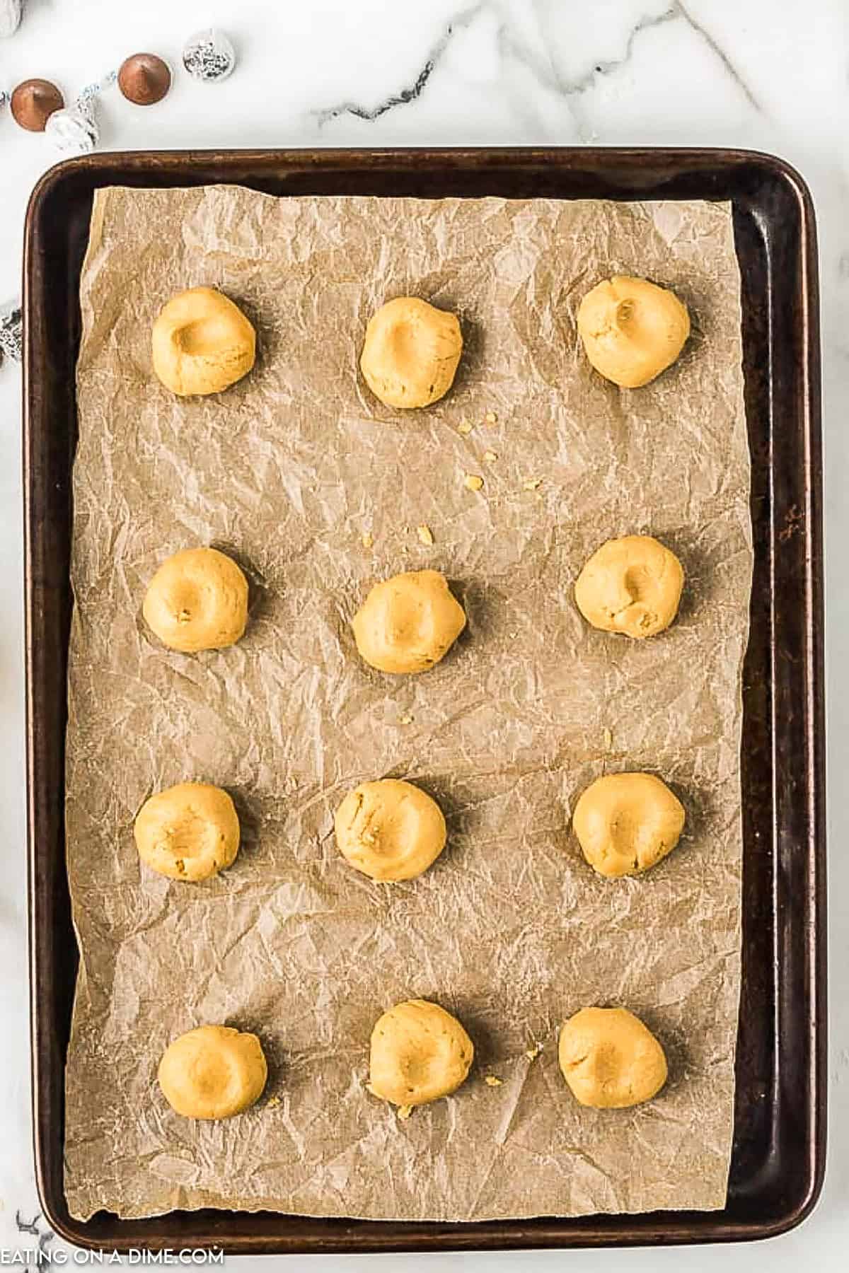 A dark baking sheet lined with parchment paper holds twelve uniform, unbaked peanut butter kiss cookie dough balls, evenly spaced in a grid pattern. The baking sheet is set on a marble countertop with a corner showing some chocolate candy in the background.