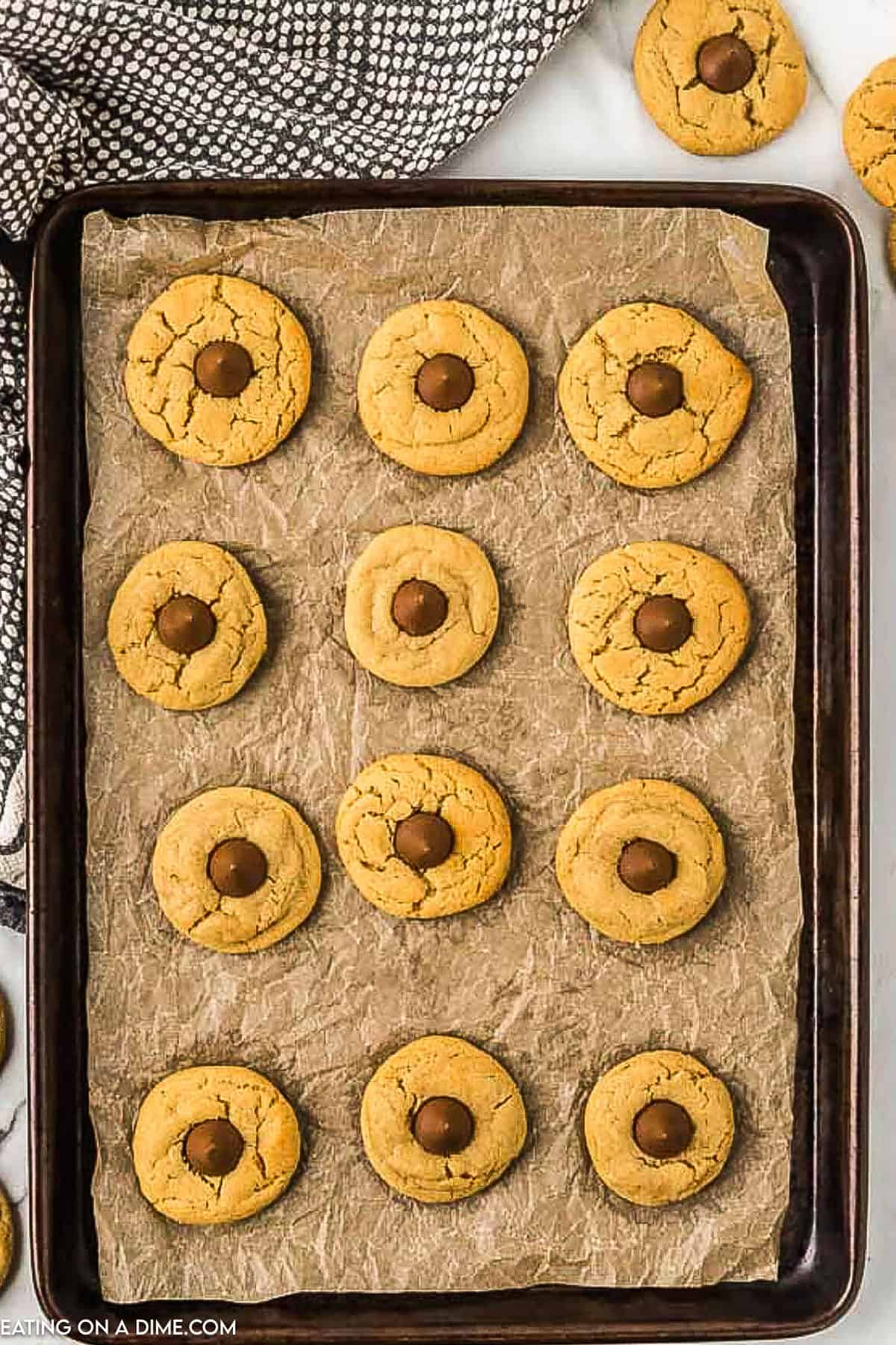 A baking sheet lined with parchment paper holds twelve freshly baked peanut butter kiss cookies, each topped with a chocolate kiss in the center. A black and white cloth is partially visible in the top left corner of the image.