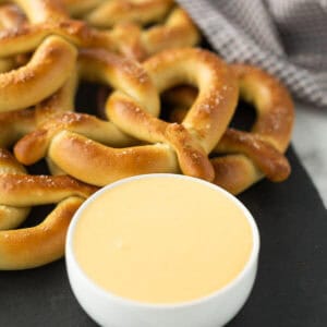 A pile of salted pretzels on a dark surface next to a small white bowl filled with cheese sauce for pretzels. A checked cloth is in the background.