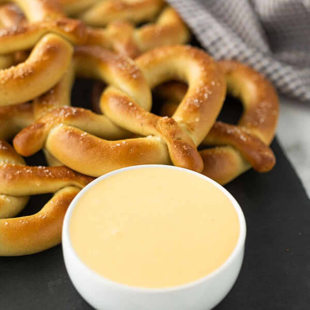 A pile of salted pretzels on a dark surface next to a small white bowl filled with cheese sauce for pretzels. A checked cloth is in the background.