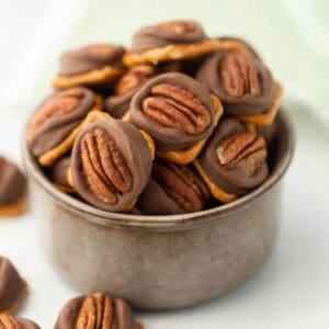 A ceramic bowl brims with chocolate-covered candies and whole pecans, echoing the sweetness of pecan Rolo pretzels. A few treats spill artfully onto the surface, while a softly blurred background hints at a green cloth.