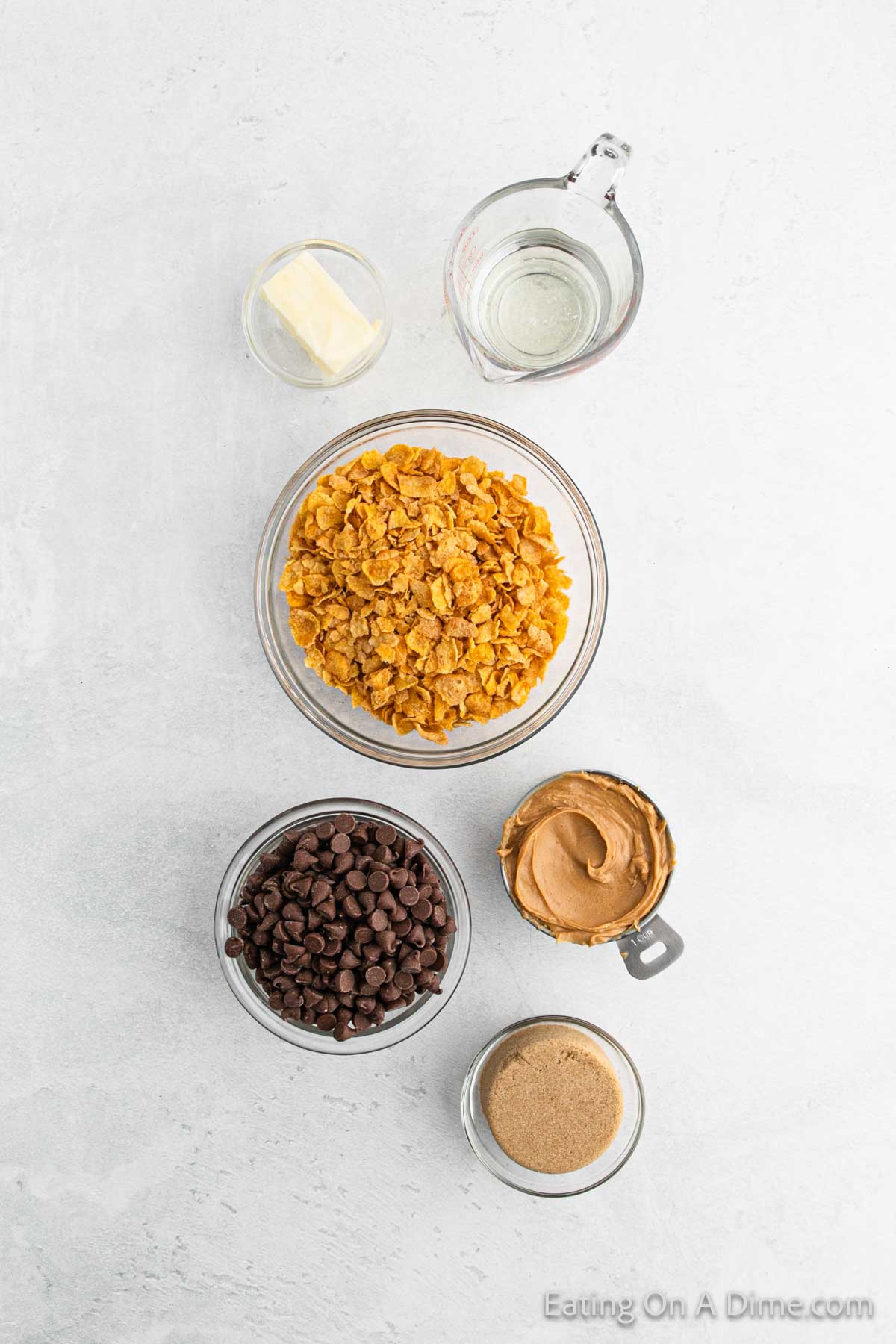 Top-down view of ingredients on a white surface for Cinnamon Cornflake Squares: a bowl of cornflakes, peanut butter in a jar, chocolate chips in a bowl, round brown sugar, butter in a small dish, and a measuring cup of clear liquid.