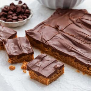 A batch of chocolate-covered peanut butter bars is laid out on parchment paper, some cut into squares resembling Cinnamon Cornflake Squares. The rest remains uncut, with a small bowl of chocolate chips visible in the background.