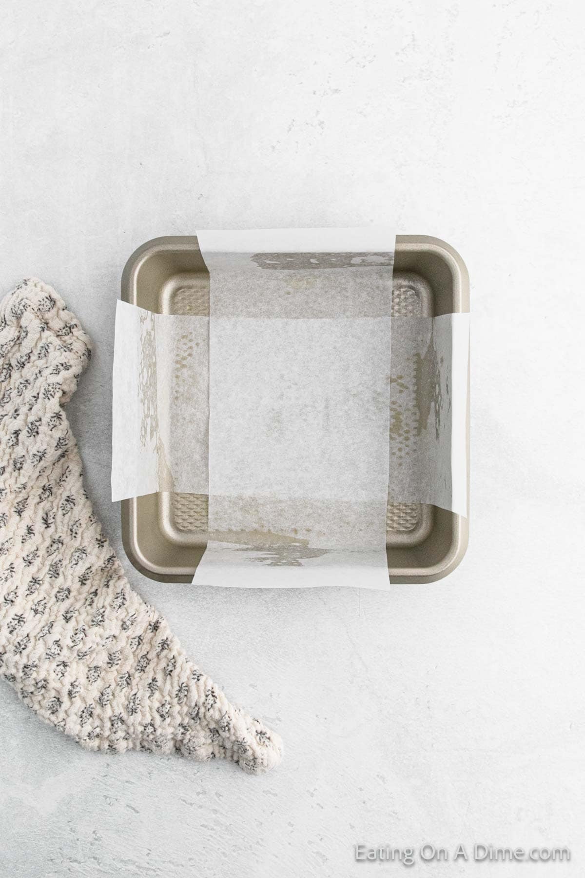 A square baking pan lined with parchment paper sits on a light gray surface, ready to transform into delicious cinnamon cornflake squares. To the left, a textured knit cloth adds a cozy touch.