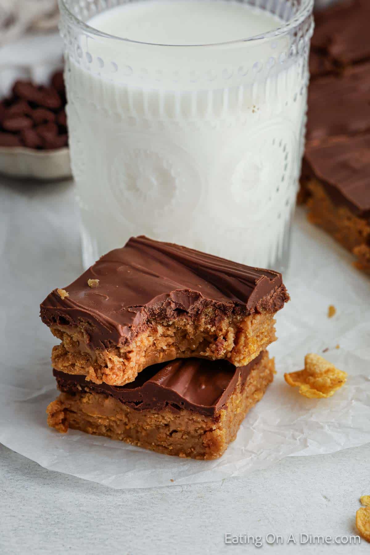 Two chocolate-covered peanut butter bars, one with a bite taken out, are stacked on parchment paper alongside cinnamon cornflake squares. Behind them is a glass of milk with an embossed pattern, and a small bowl of chocolate chips peeks into view.