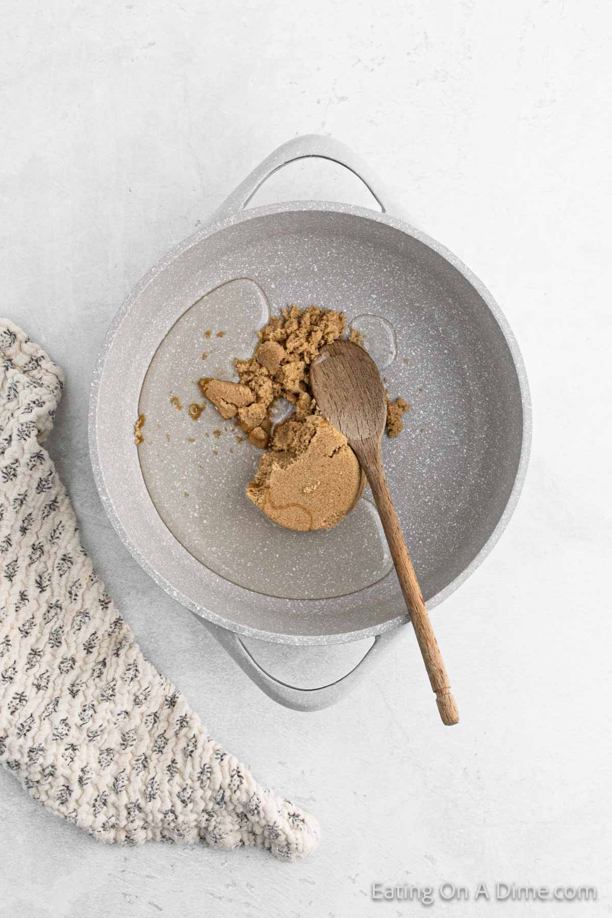 A pot with brown sugar and a wooden spoon inside rests on a light-colored surface, evoking the cozy scent of Cinnamon Cornflake Squares. A textured beige cloth lies beside the pot, completing the warm and inviting scene.