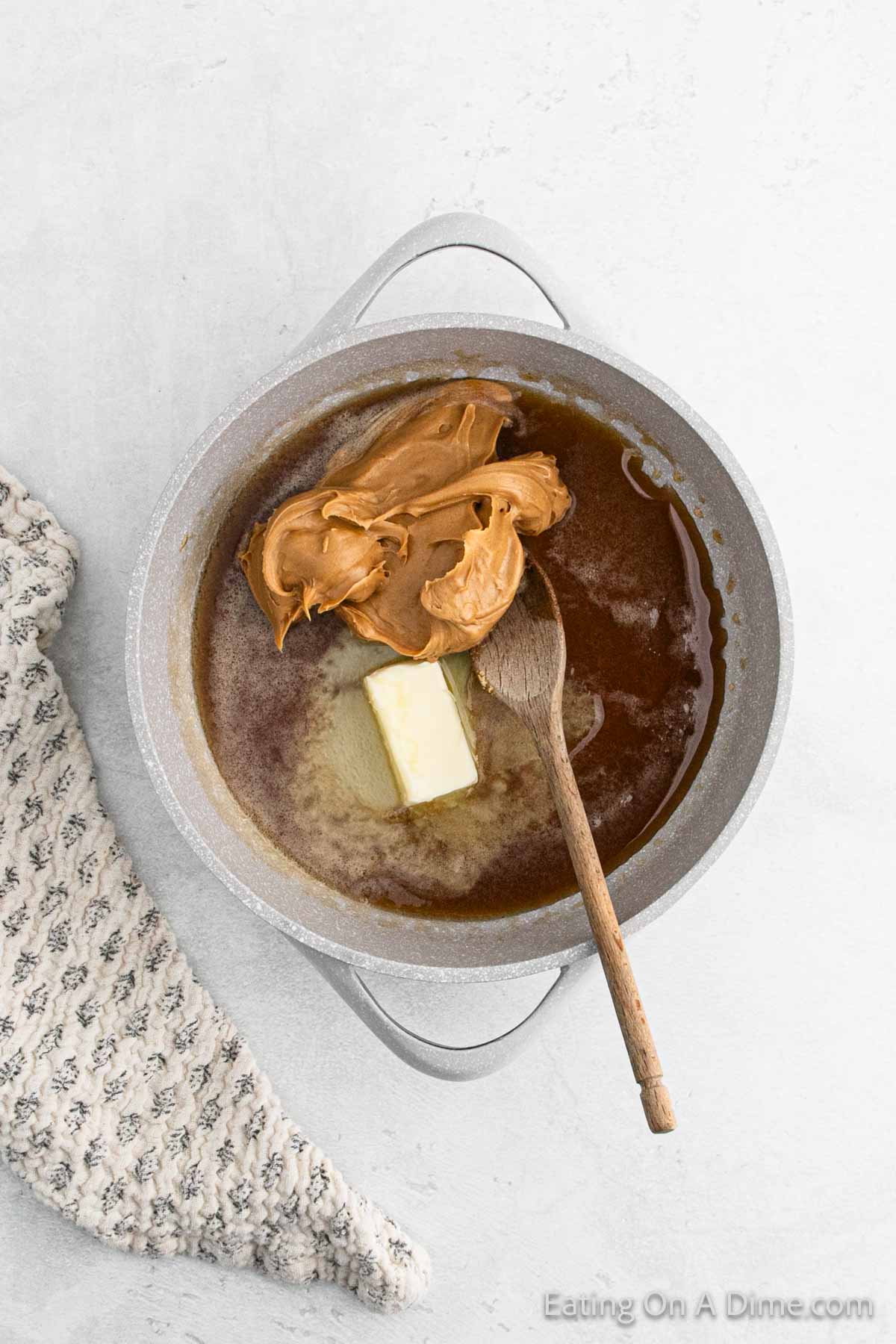 A pot on a light surface with melted butter, brown sugar, and peanut butter, perfect for creating Cinnamon Cornflake Squares. A wooden spoon rests inside, ready for mixing. A textured cloth is placed beside the pot.