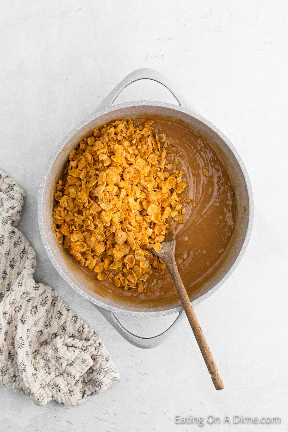 A pot filled with a creamy, brown batter and crushed cornflakes awaits transformation into Cinnamon Cornflake Squares. A wooden spoon rests beside the inviting concoction, while a patterned cloth lies nearby on a light gray surface.