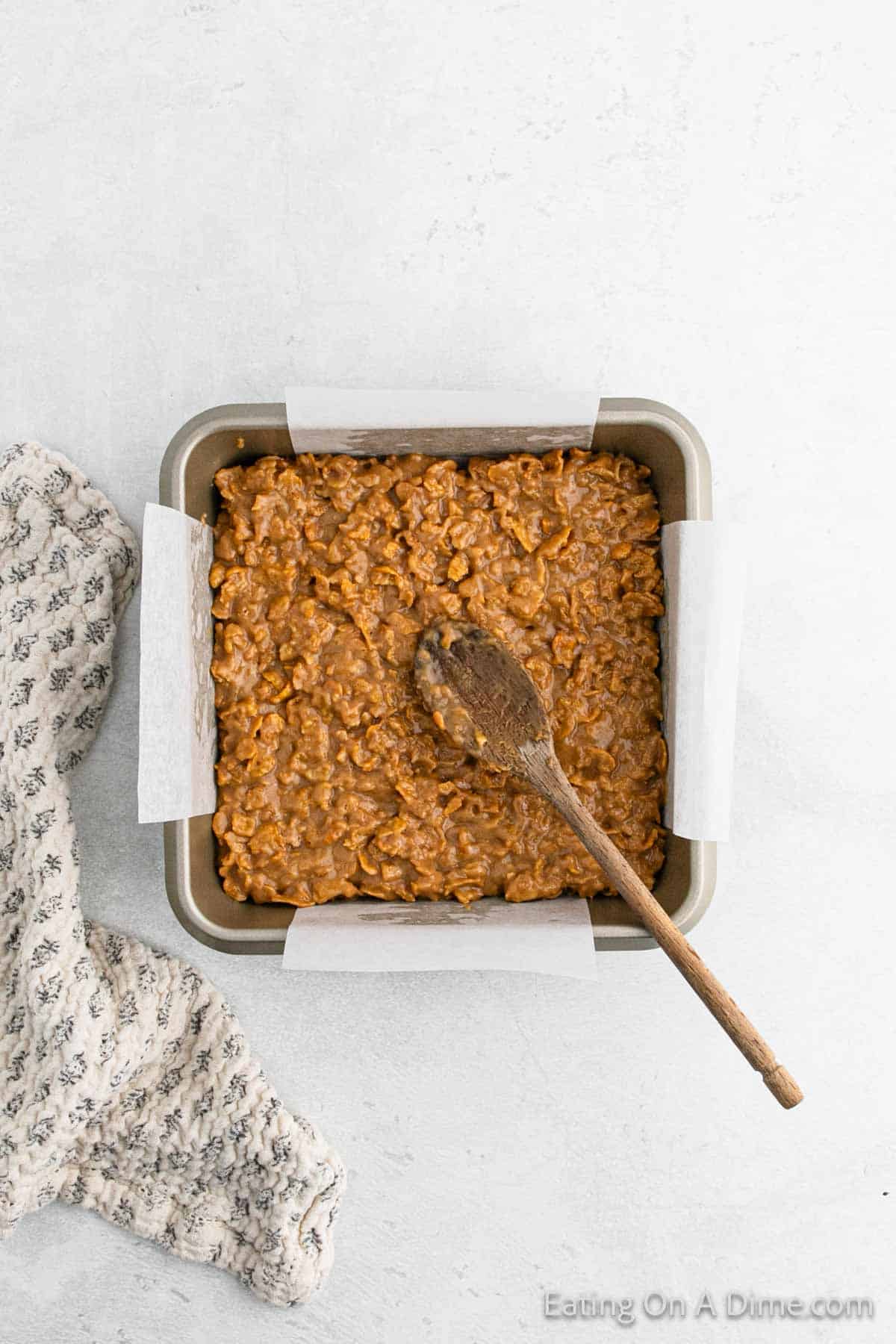 A square baking pan filled with cinnamon-infused batter mixture, with a wooden spoon resting on top. The pan is lined with parchment paper, ready to bake delicious Cinnamon Cornflake Squares. A patterned cloth is draped beside the pan on a light surface.