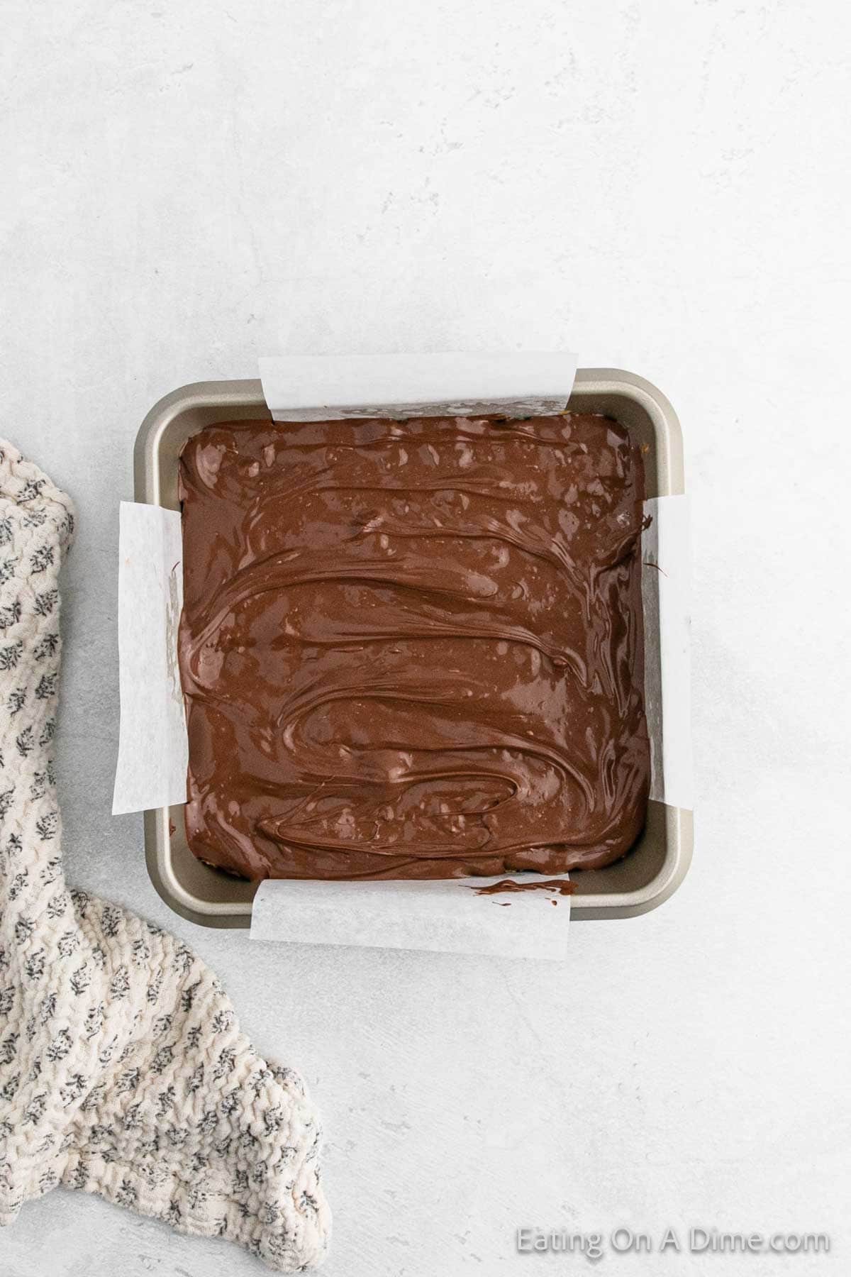 A square baking pan lined with parchment paper is filled with thick, creamy chocolate batter, promising the decadent crunch of Cinnamon Cornflake Squares. The pan rests on a light surface, partially covered by a textured cloth.