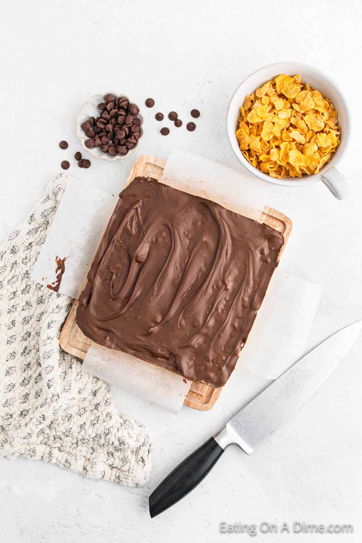 A square chocolate-covered dessert, known as a Cinnamon Cornflake Square, rests on parchment paper atop a wooden board. Nearby lie a bowl of cornflakes, chocolate chips, a knife, and a patterned cloth.