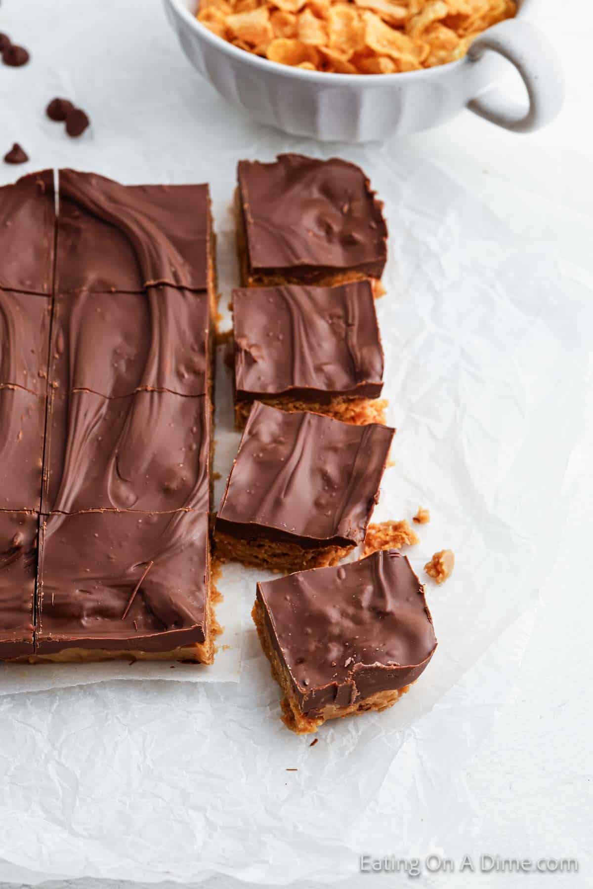 Cinnamon Cornflake Squares of chocolate peanut butter bars rest on parchment paper, with a bowl of cornflakes in the background.