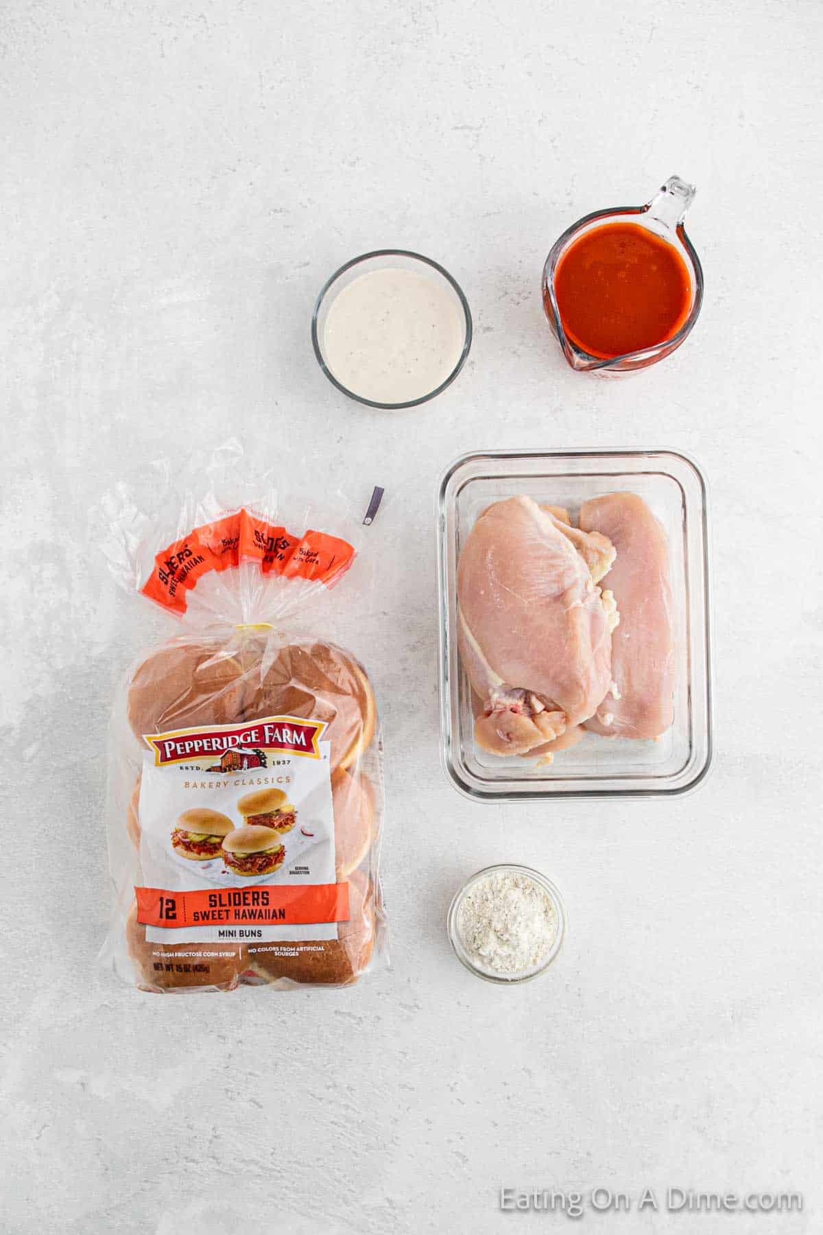 An arrangement of cooking ingredients suggests a delightful meal in the making: a package of sweet Hawaiian slider buns, raw chicken breasts ready for the crock pot, Parmesan cheese, a vibrant red sauce, and ranch dressing—all set for an irresistible Buffalo Chicken Sliders recipe.