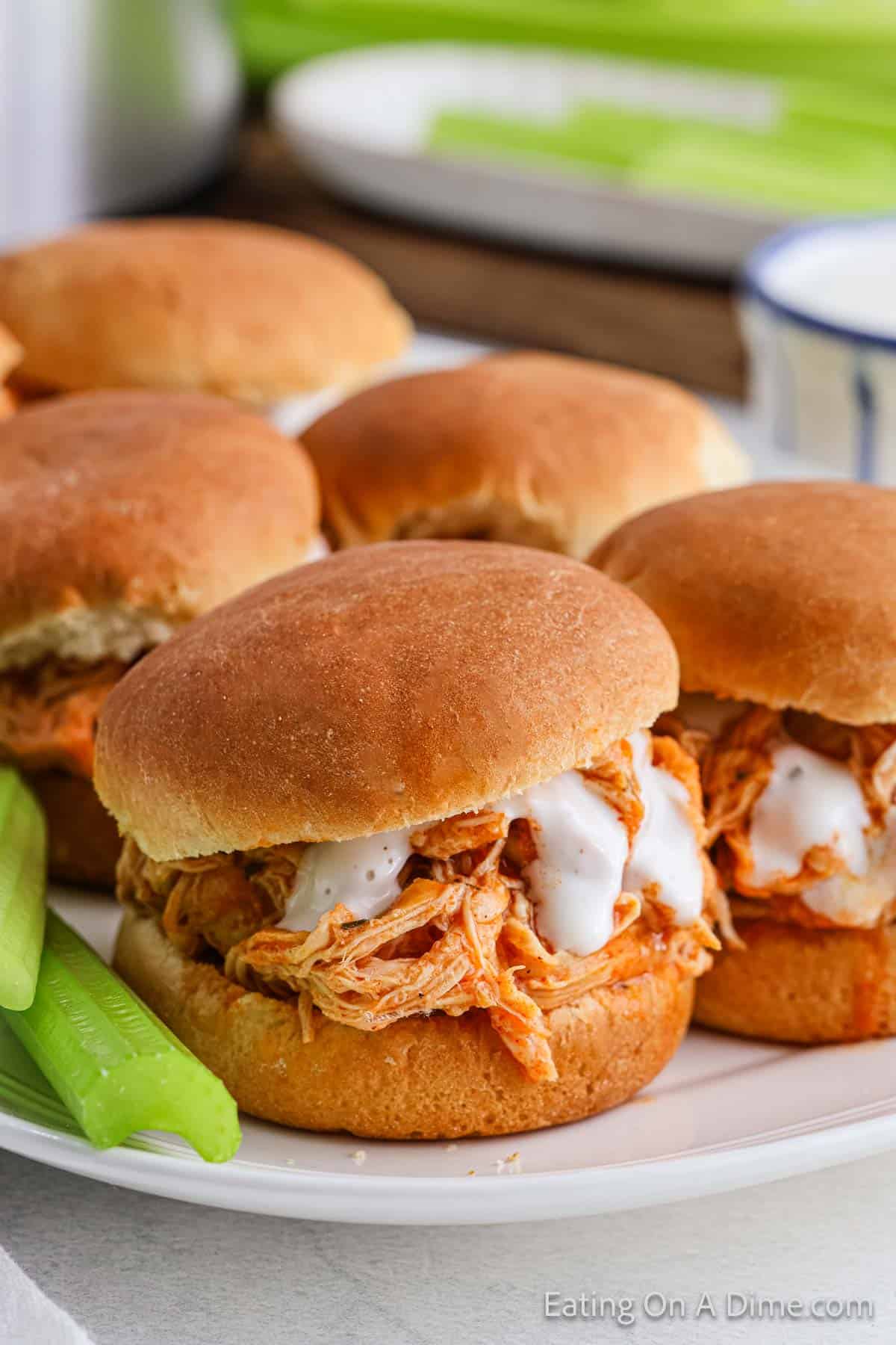 This Crock Pot Buffalo Chicken Sliders recipe features a mouthwatering close-up of four sliders topped with tangy sauce, nestled on fresh buns. Crisp green celery sticks are thoughtfully placed alongside, ready to complement the spicy flavor explosion.
