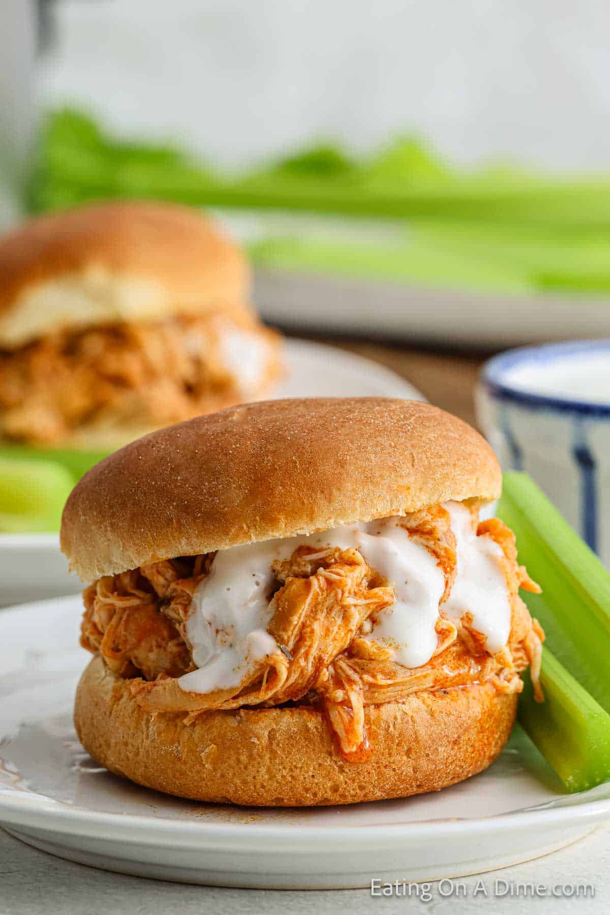 A close-up of a Crock Pot Buffalo Chicken Slider topped with creamy white sauce on a bun, served on a white plate. A stalk of celery rests next to the sandwich. Another slider and celery are blurred in the background.