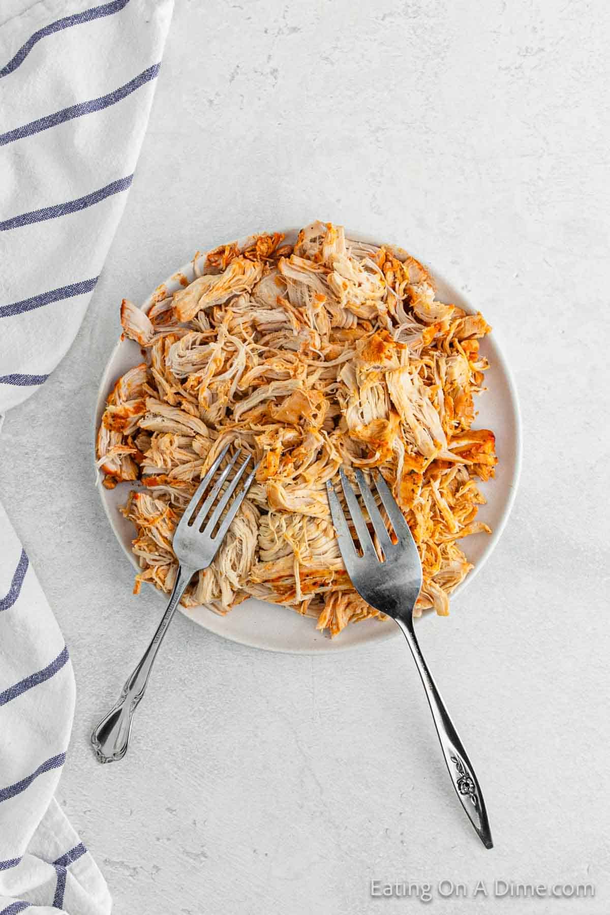 A plate of shredded chicken, perfect for Crock Pot Buffalo Chicken Sliders, is seasoned with spices and rests on a light surface. Two forks wait eagerly on the plate for serving. A blue and white striped cloth is partially visible to the left.