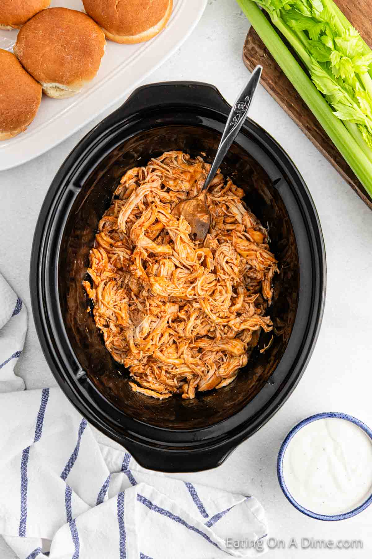 A crock pot buffalo chicken sliders recipe features shredded buffalo chicken in a black slow cooker with a silver spoon. Nearby, find sandwich buns, crisp celery stalks, a bowl of ranch dressing, and a striped cloth elegantly placed on the white surface.