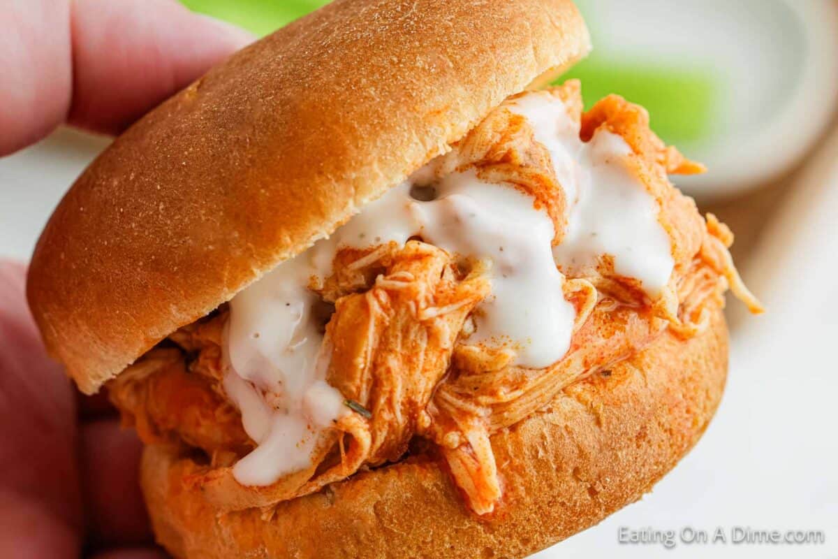 A close-up of a Crock Pot Buffalo Chicken Slider on a bun, topped with creamy sauce. The sandwich is being held in a hand, against a blurred green background.