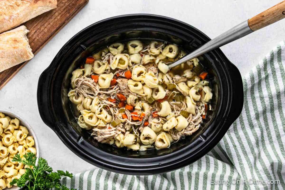 A black crockpot warms a hearty chicken tortellini soup with shredded chicken, carrots, and a flavorful broth. A silver ladle rests inside, ready to serve. Fresh bread and uncooked tortellini sit nearby on a striped cloth, completing the inviting scene.