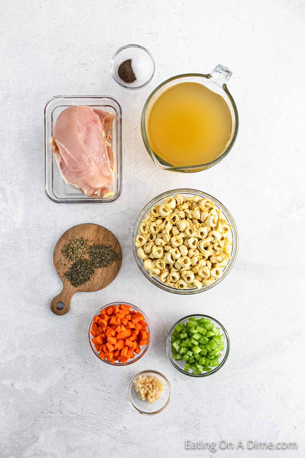 Ingredients for a crockpot chicken tortellini soup are laid out on a white surface, featuring chicken breast, broth, tortellini pasta, chopped carrots and celery, minced garlic, and herbs on a wooden board. Don't forget the salt and pepper for seasoning this comforting dish!