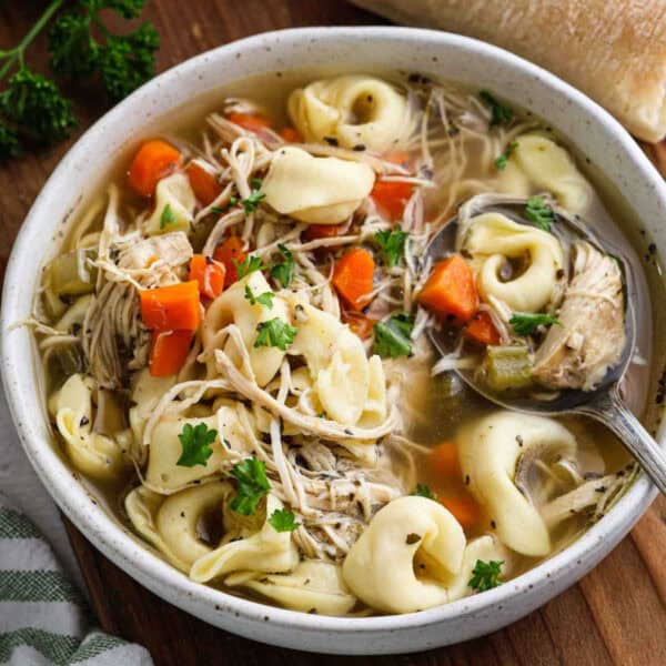 A bowl of crockpot chicken tortellini soup brimming with shredded chicken, carrots, celery, and herbs. A spoon rests inside the bowl, with a piece of bread partially visible in the background. Fresh parsley garnishes this comforting dish.