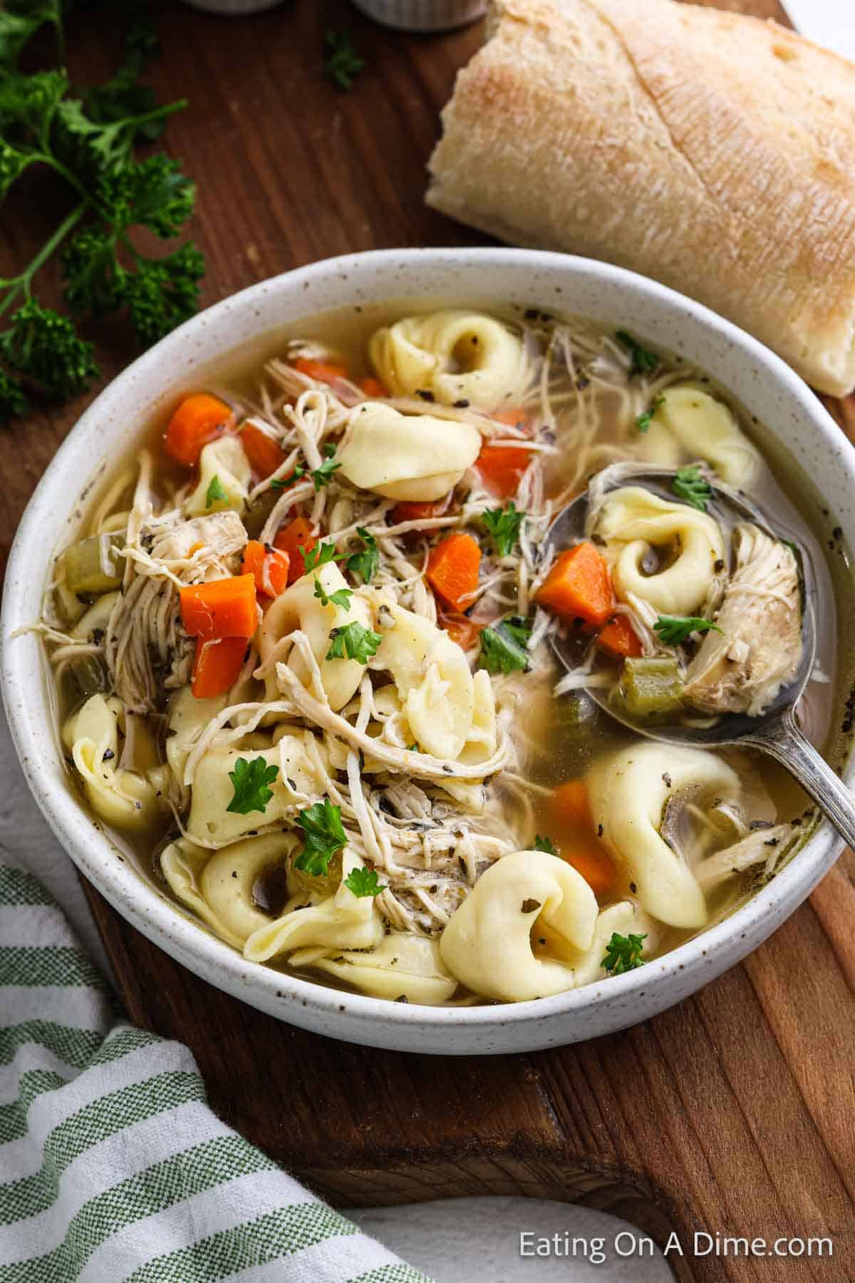 A bowl of crockpot chicken tortellini soup filled with tender pasta, shredded chicken, carrots, and celery, garnished with parsley. A slice of crusty bread and a sprig of fresh parsley sit beside the bowl. A green and white striped cloth is partially visible.