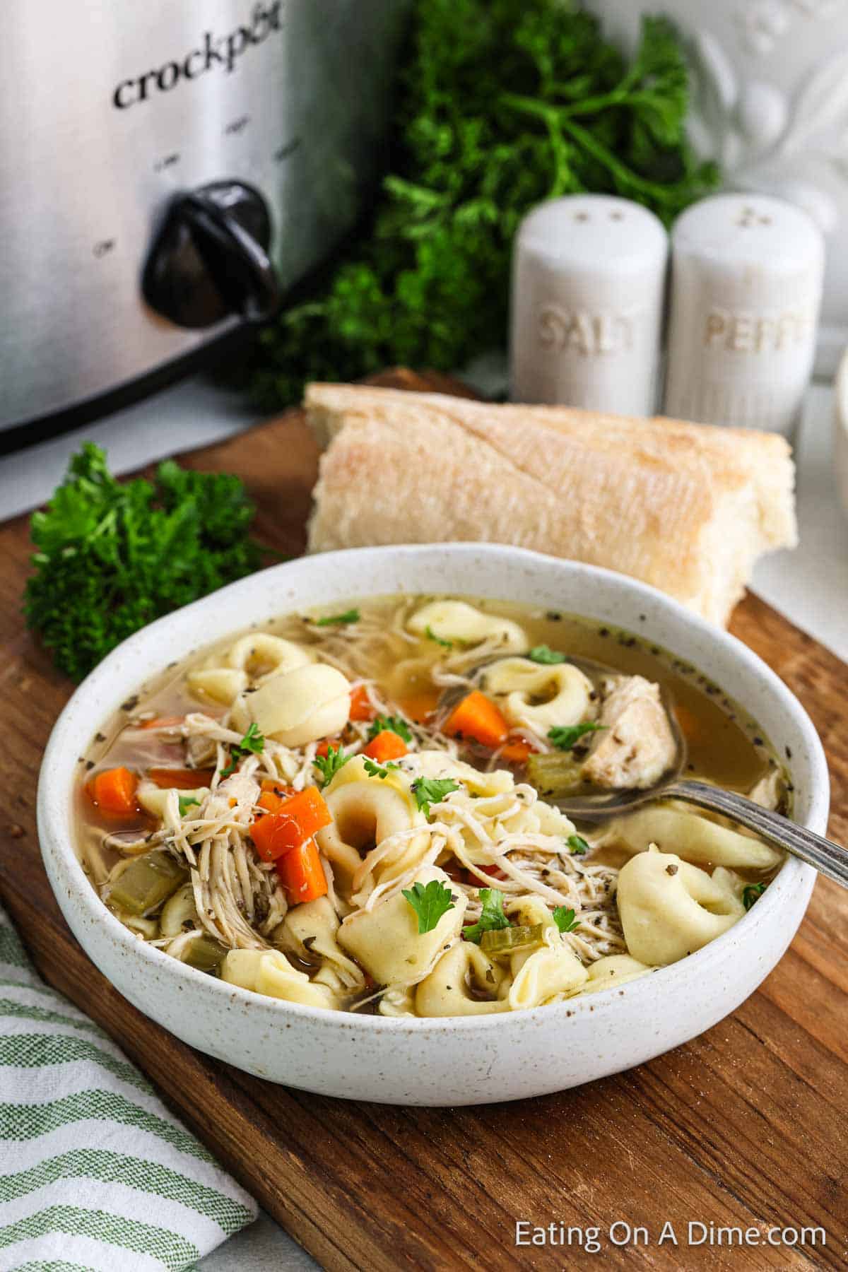 A bowl of crockpot chicken tortellini soup with carrots and herbs sits on a wooden board. A piece of bread and a sprig of greenery rest beside it, while a salt and pepper shaker can be seen alongside the slow cooker in the background.