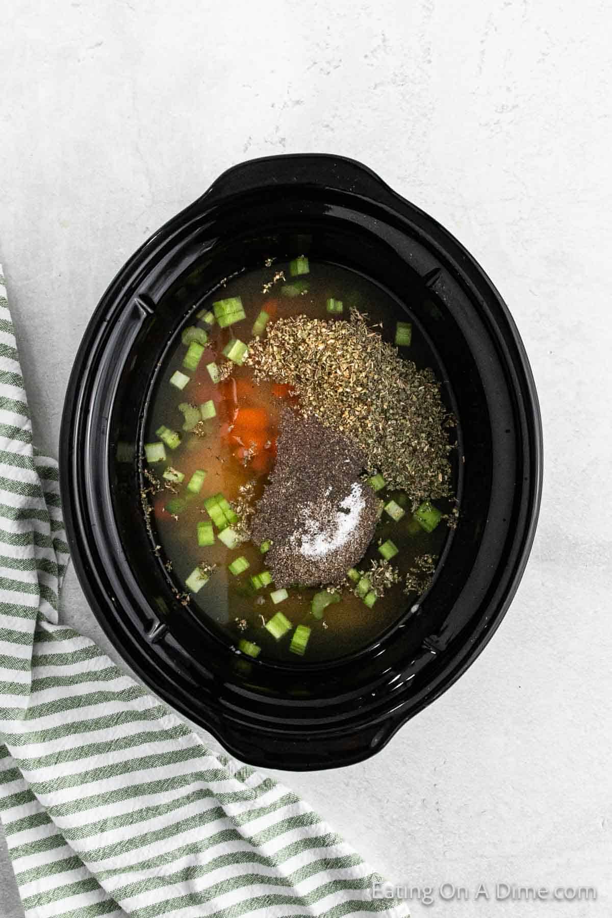 A black slow cooker brimming with a colorful mixture of diced vegetables, spices, and broth for a hearty crockpot chicken tortellini soup. Ingredients include chopped celery, herbs, and black pepper. A green and white striped cloth rests beside the cooker on a light surface.