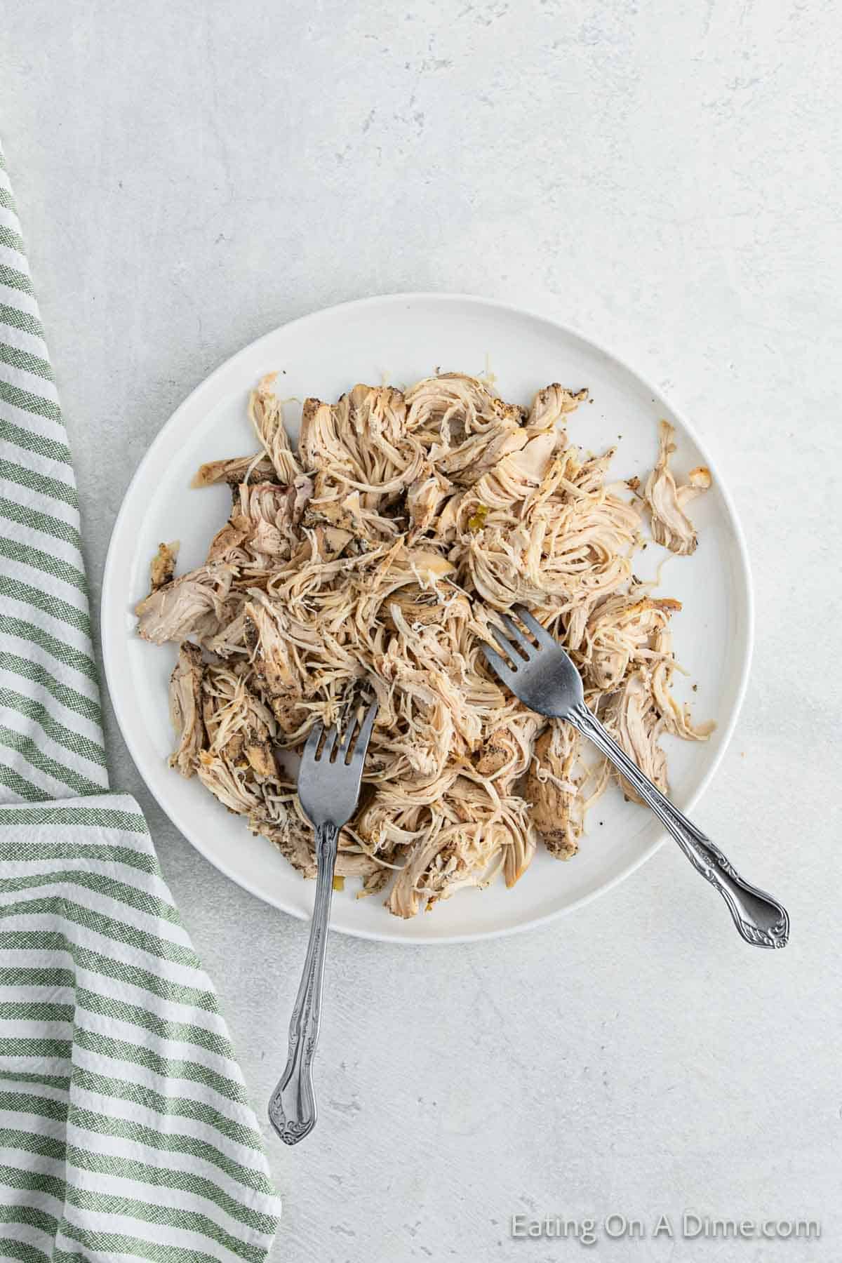 A white plate filled with shredded crockpot chicken, garnished with herbs. Two forks rest on the plate. A green and white striped cloth is partially visible on the left side. The surface is light gray, reminiscent of a cozy kitchen scene ready for tortellini soup inspiration.