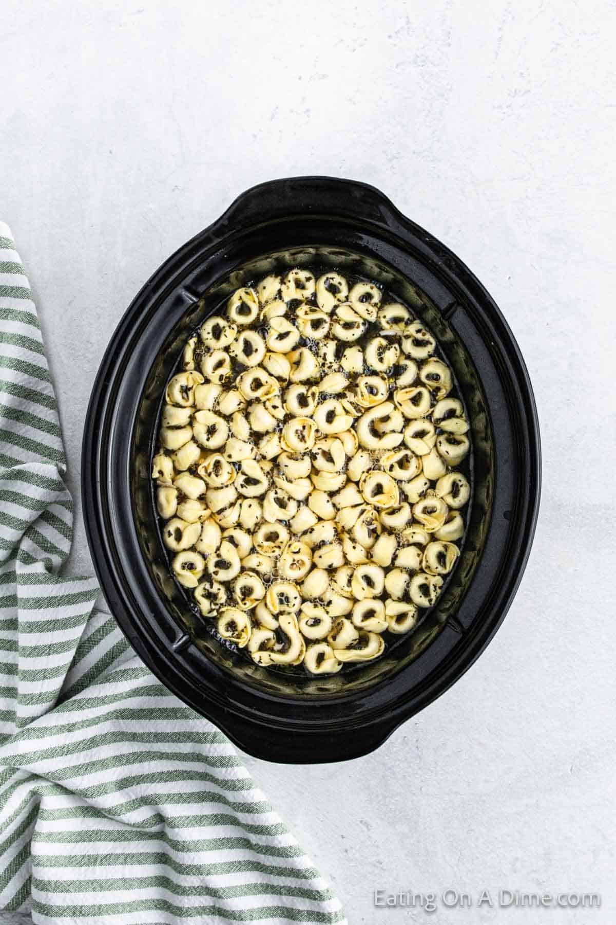 A black slow cooker filled with delicious tortellini, seasoned with herbs, sits on a light gray surface. This savory dish, reminiscent of a hearty crockpot chicken tortellini soup, is beautifully accented by a striped green and white cloth draped beside it.