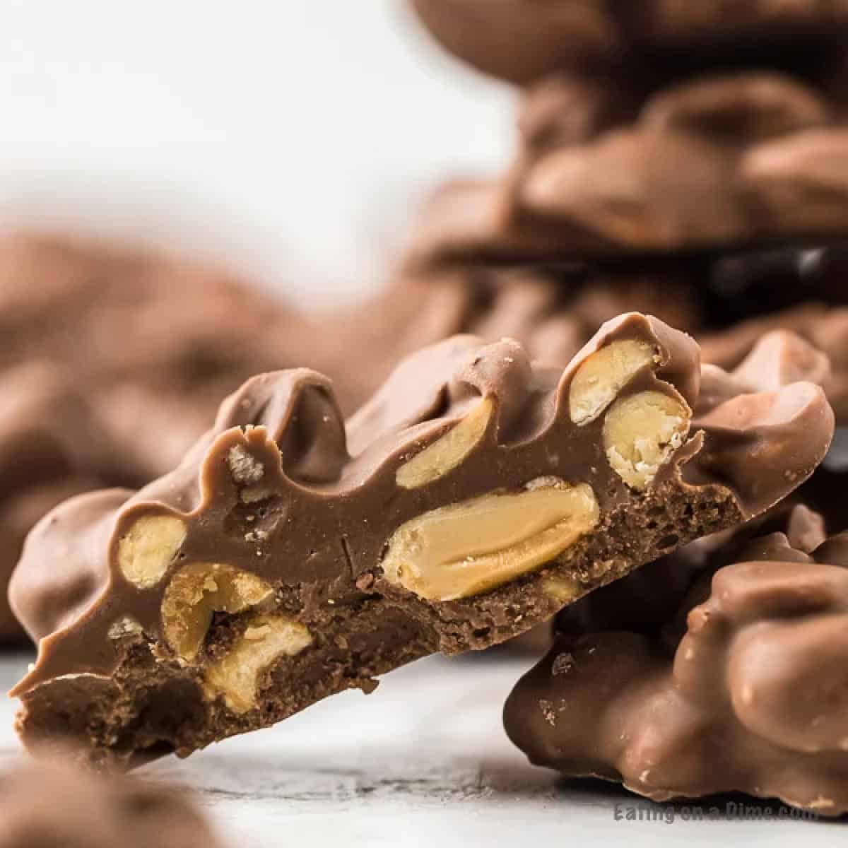 Close-up of a stack of Crockpot Peanut Clusters. The front cluster is broken in half, revealing whole peanuts embedded in rich, creamy chocolate. The background shows additional clusters, slightly blurred, emphasizing texture and detail in the treats.