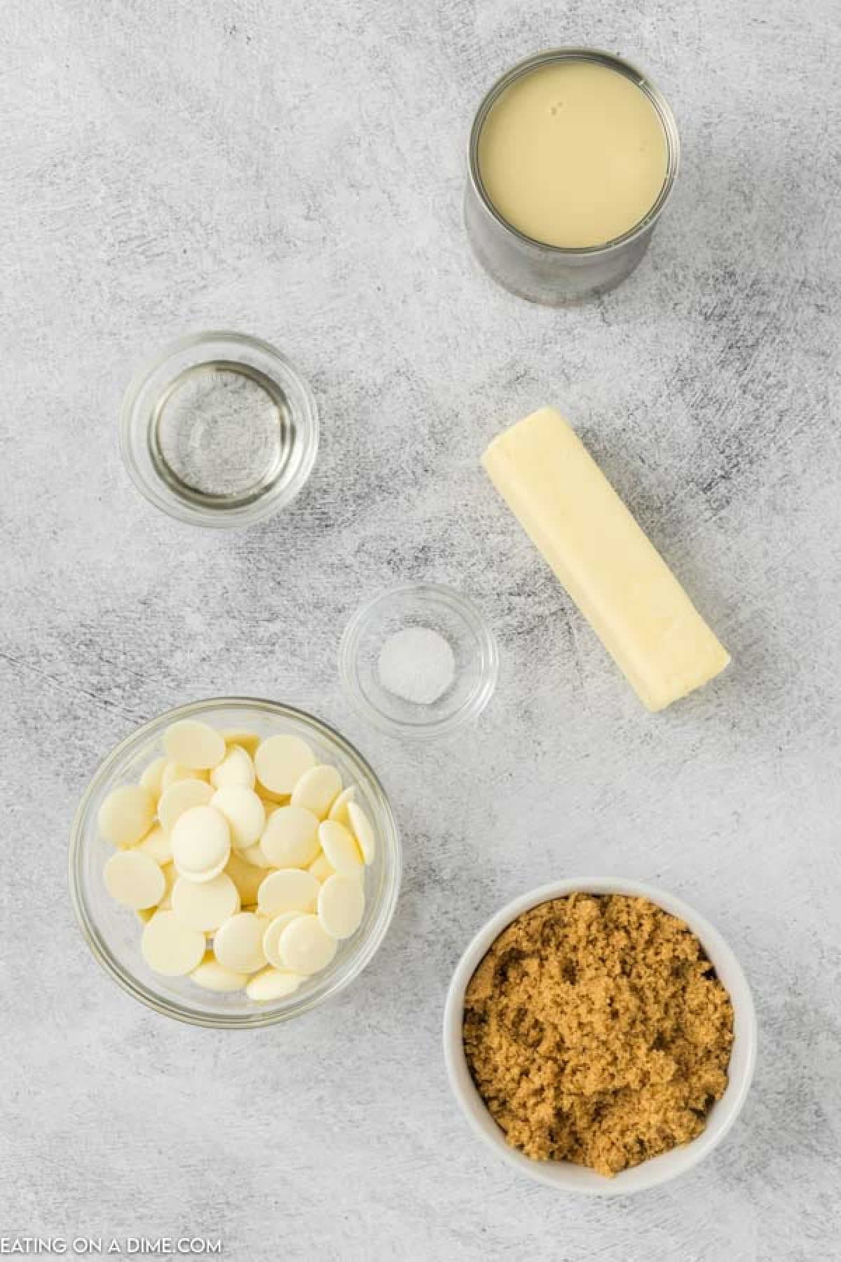 Ingredients for an easy caramel fudge recipe on a countertop: a stick of butter, a can of sweetened condensed milk, a bowl of white chocolate chips, a bowl of brown sugar, a small bowl of salt, and a small bowl of vanilla extract.