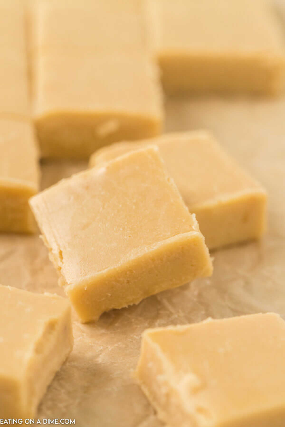 Close-up of several square pieces of creamy vanilla fudge arranged on parchment paper. The fudge, made from an easy caramel fudge recipe, has a smooth, light beige texture and appears freshly cut. The background is softly out of focus.