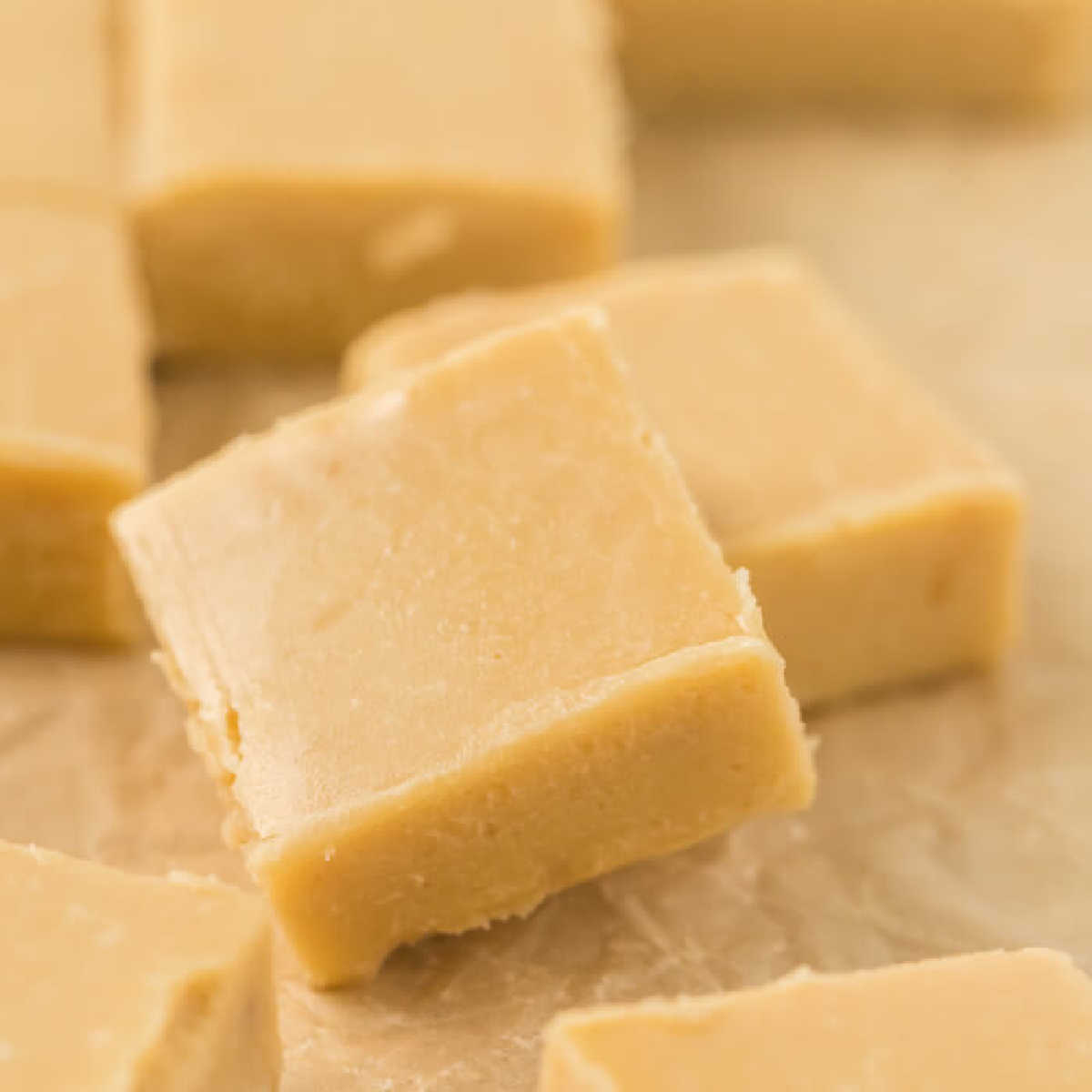 Close-up of several pieces of creamy, beige fudge. The squares of fudge are arranged on a light-colored surface, showcasing a slightly soft and smooth texture that suggests an easy caramel fudge recipe, freshly made.