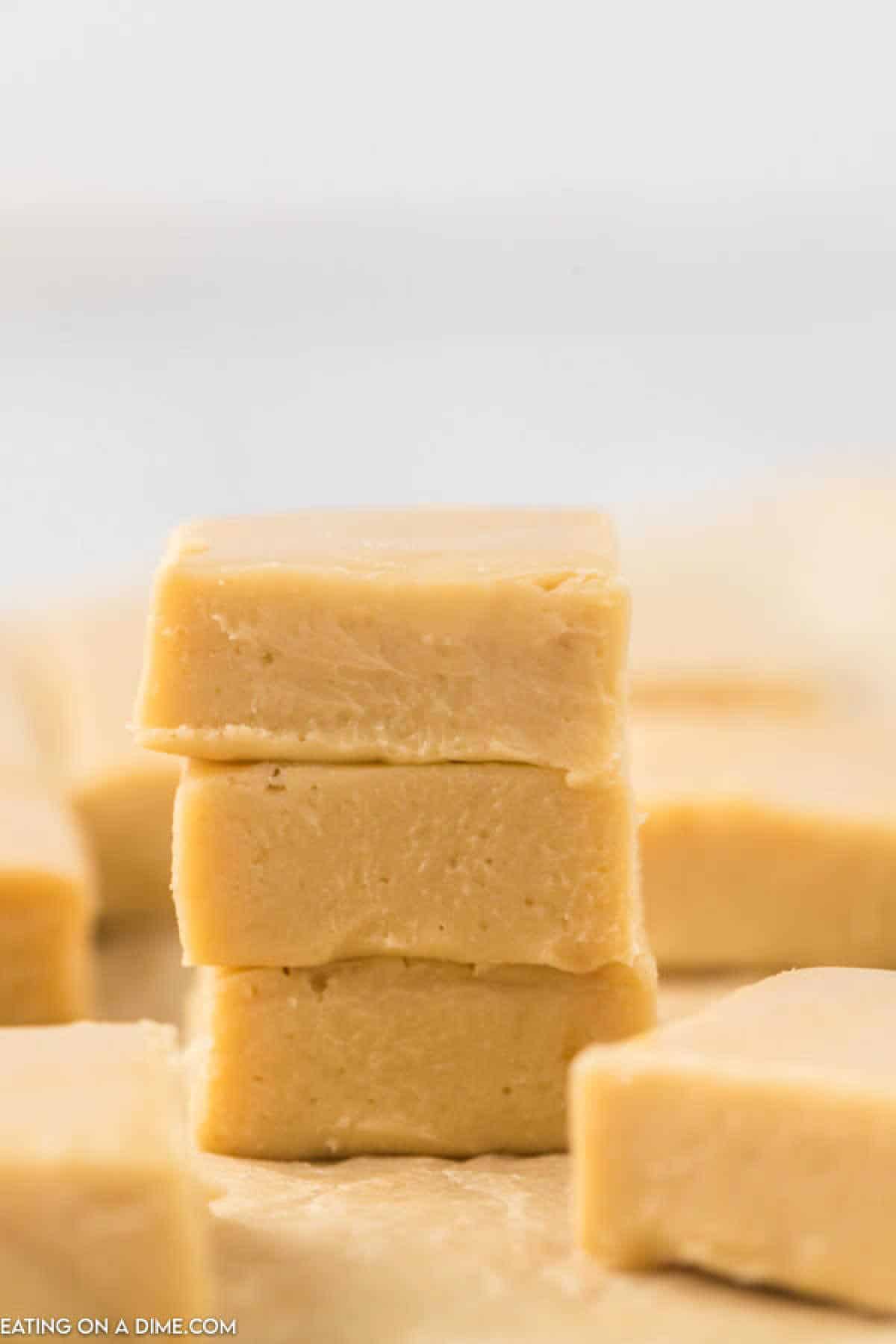 A close-up photo of a stack of three creamy, rectangular pieces of homemade peanut butter fudge from an easy caramel fudge recipe. The fudge has a smooth, slightly glossy texture with small, visible imperfections. Additional pieces of fudge are blurred in the background.