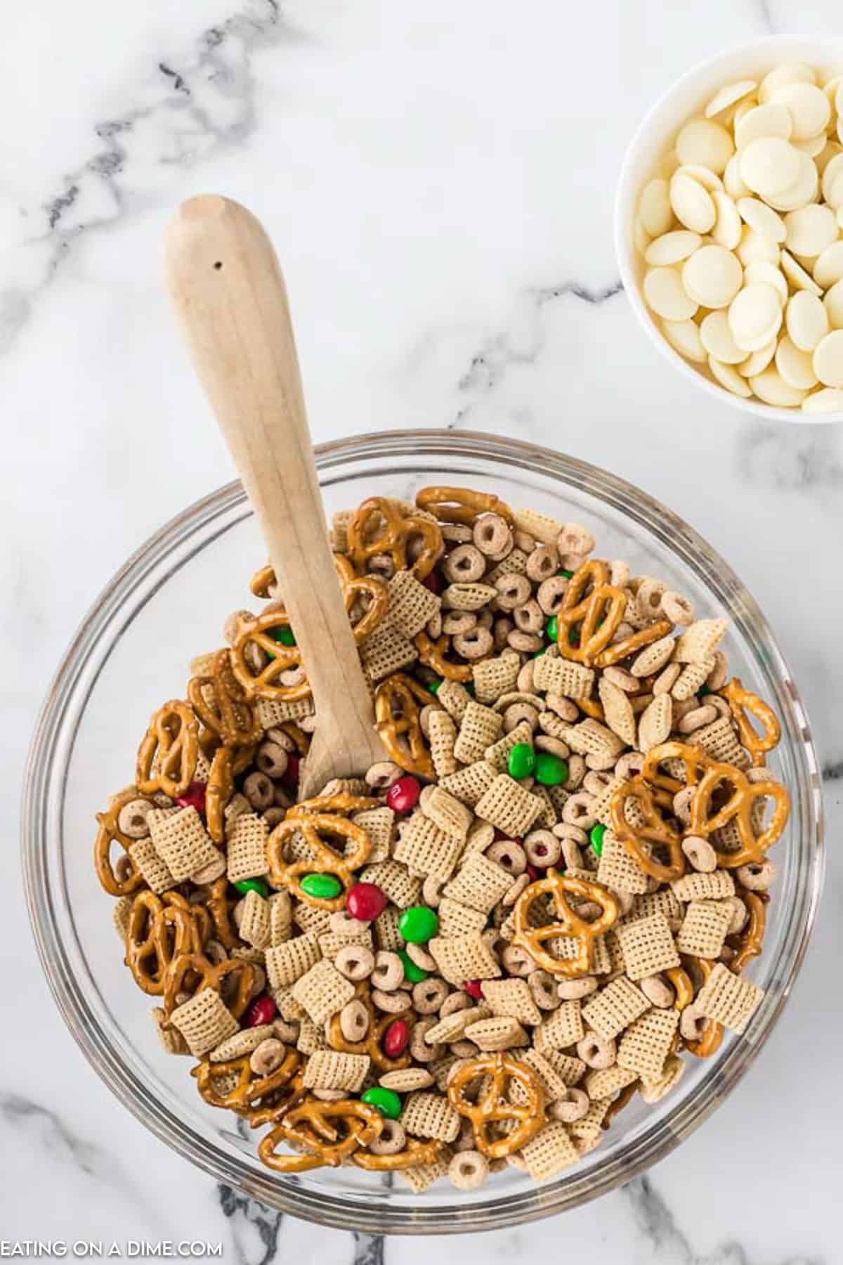 A glass bowl filled with a festive Christmas Chex Mix of pretzels, Chex cereal, Cheerios, and red and green candies, with a wooden spoon placed inside. Next to it is a smaller bowl containing white chocolate chips, all set on a marble countertop.