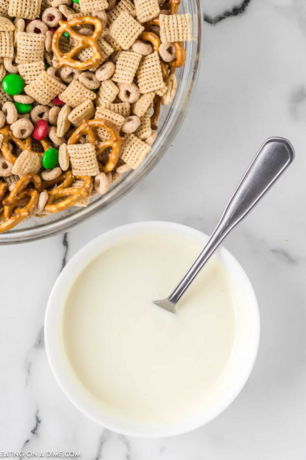 A large glass bowl filled with a festive mix of pretzels, Chex cereal, Cheerios, and colorful M&M candies sits on a marble countertop. This Christmas Chex Mix is complemented by a smaller white bowl containing creamy melted chocolate with a spoon resting inside.