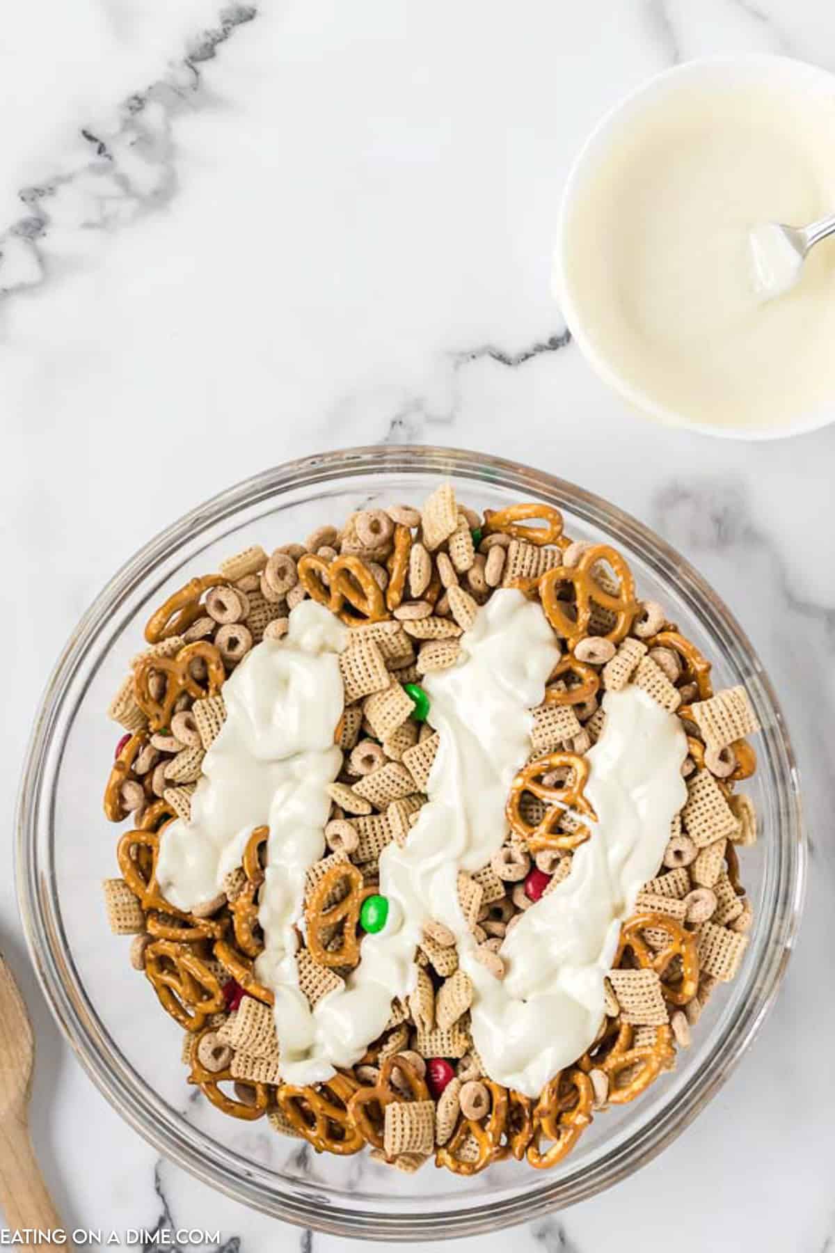 A large glass bowl filled with Christmas Chex Mix features cereals, pretzels, and colorful candy-coated chocolates. Drizzled on top is a white coating, possibly melted white chocolate. Next to the bowl is a smaller bowl containing the same white coating and a spoon.