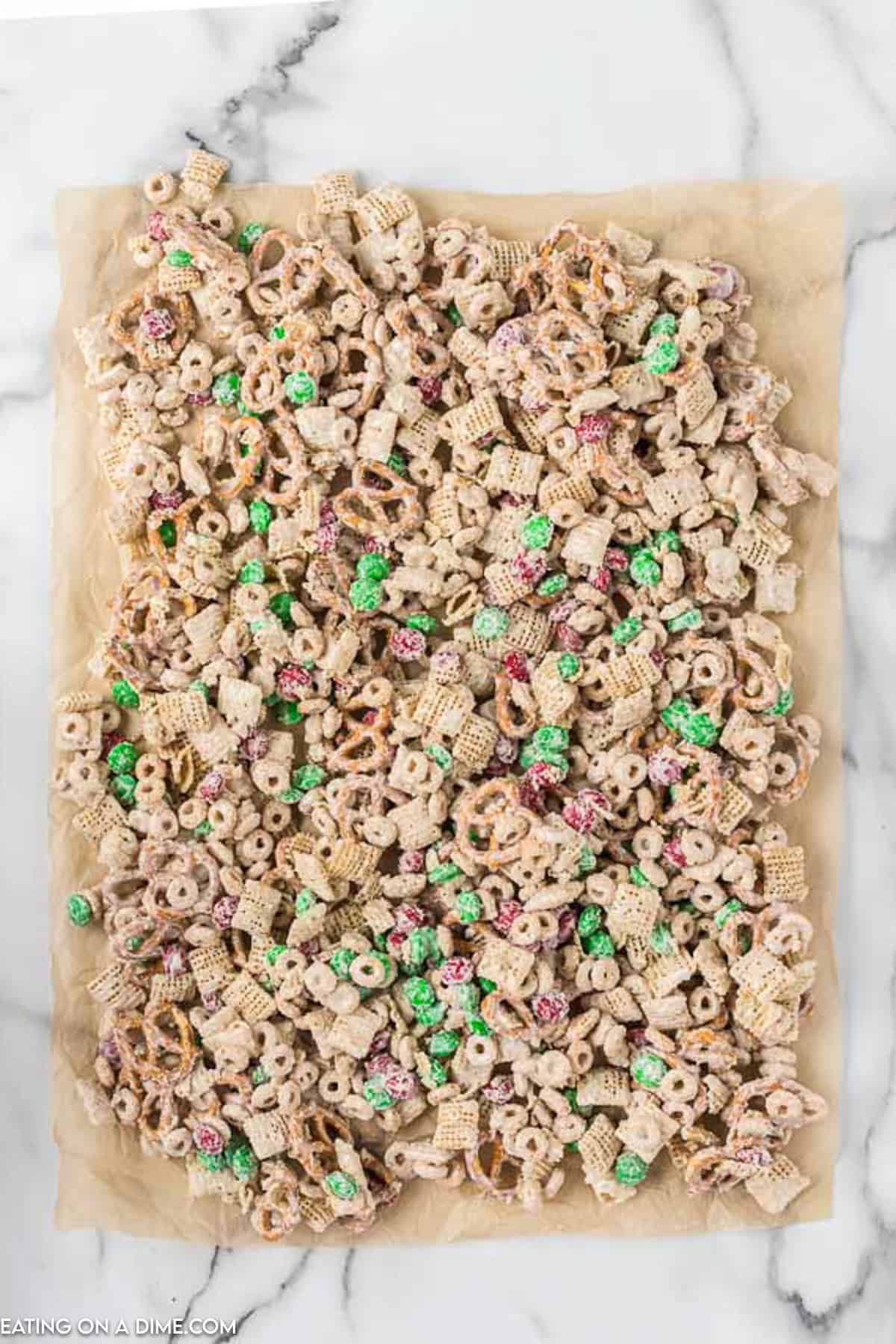 An overhead view of Christmas Chex Mix, commonly known as Reindeer Chow. It features a blend of Chex cereal, pretzels, Cheerios, and M&M candies, all coated in a white, sugary glaze, spread out over a sheet of parchment paper on a marble surface.