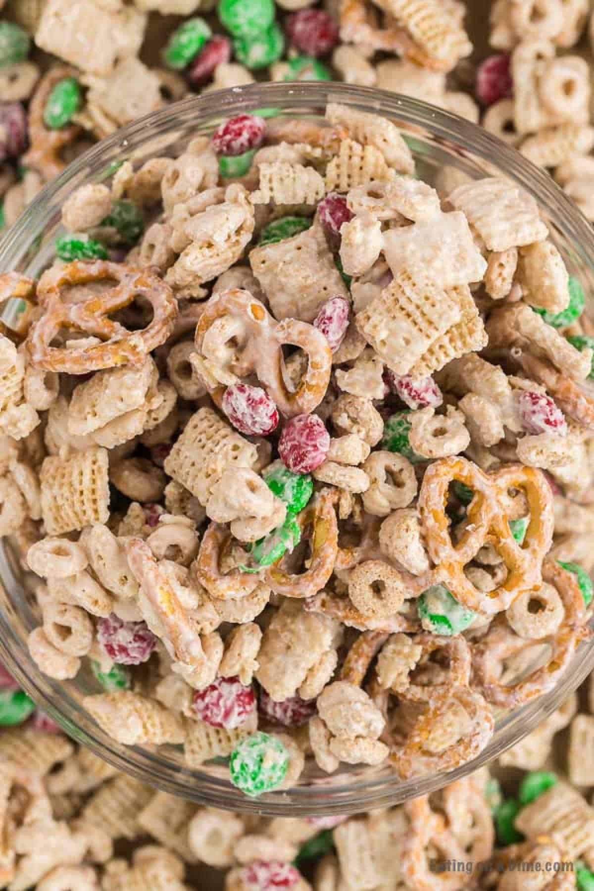 A close-up of a bowl filled with Christmas Chex Mix comprising cereal squares, pretzels, green and red M&M's, and other bite-sized treats, all coated in a white, creamy coating. The mix appears festive with bright colors and a variety of textures.