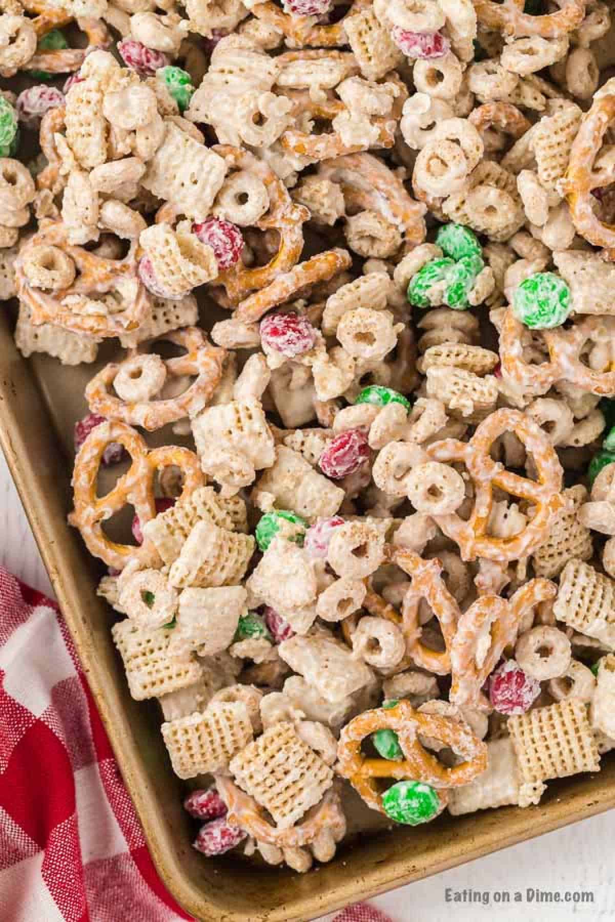 A close-up of a festive Christmas Chex Mix on a baking tray. The mix includes pretzels, Chex cereal, Cheerios, red and green M&Ms, and bits of other cereal, all coated in a white sugary glaze. A red and white checkered cloth is partially visible on the side.