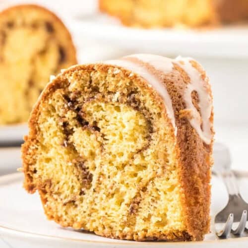 A slice of coffee cake with a marbled cinnamon swirl is topped with a light glaze, reminiscent of a homemade Cinnamon Bundt Cake recipe. It sits on a white plate, fork beside it, while another slice rests in the background, slightly out of focus.