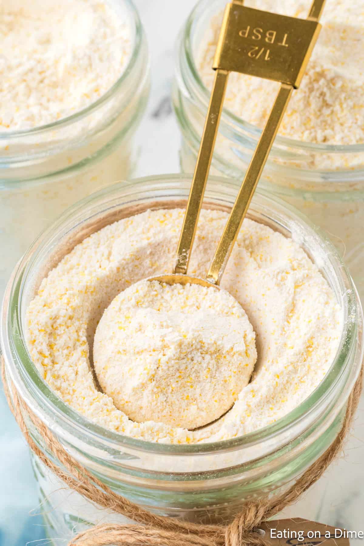 A close-up of a glass jar filled with cornmeal, a crucial ingredient for any homemade hummus recipe. A brass measuring spoon marked "½ tbsp" rests inside, ready for action. In the background, more jars await their turn, one adorned with twine around its neck for a touch of charm.